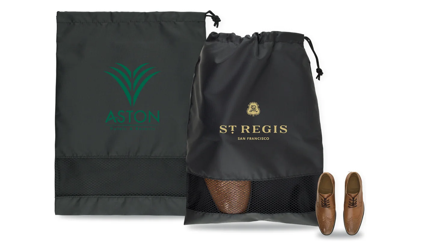Two custom printed black drawstring shoe bags with a mesh window, one with a hotel logo, beside a pair of dress shoes